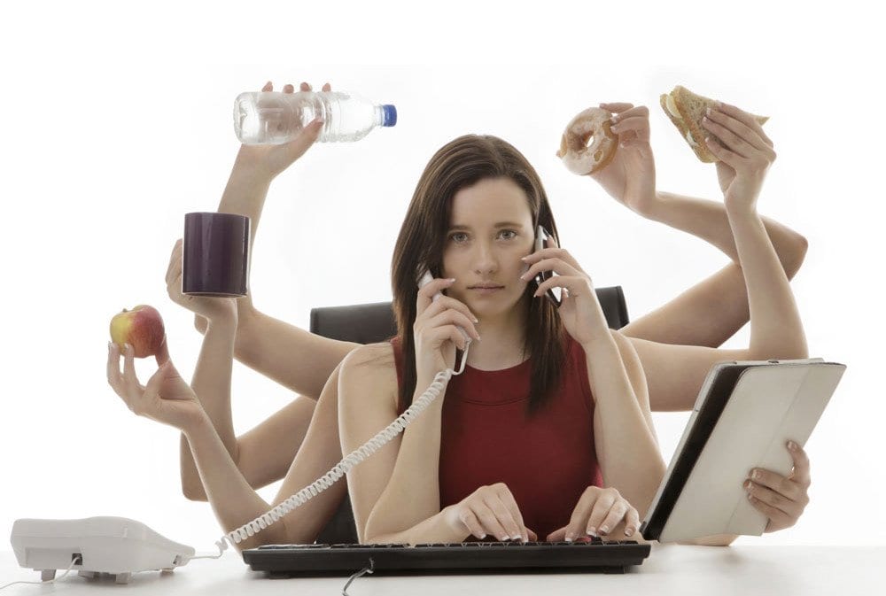 A woman in a red dress sitting at a desk in front of her computer.  3 pairs of arms are coming out form behind her body showing all the tasks she needs to do. 