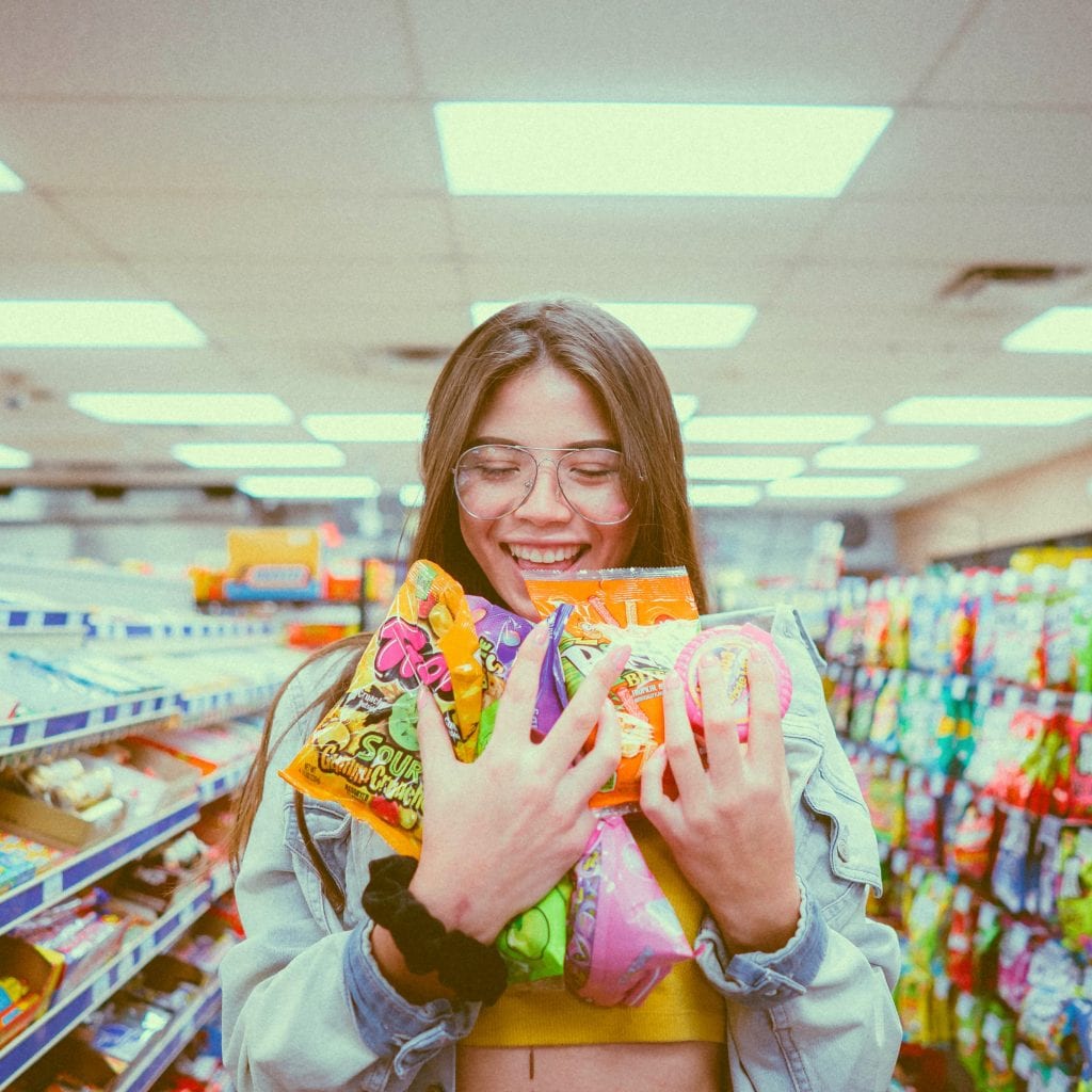 A girl in a shopping aisle with an armfull of junk food.