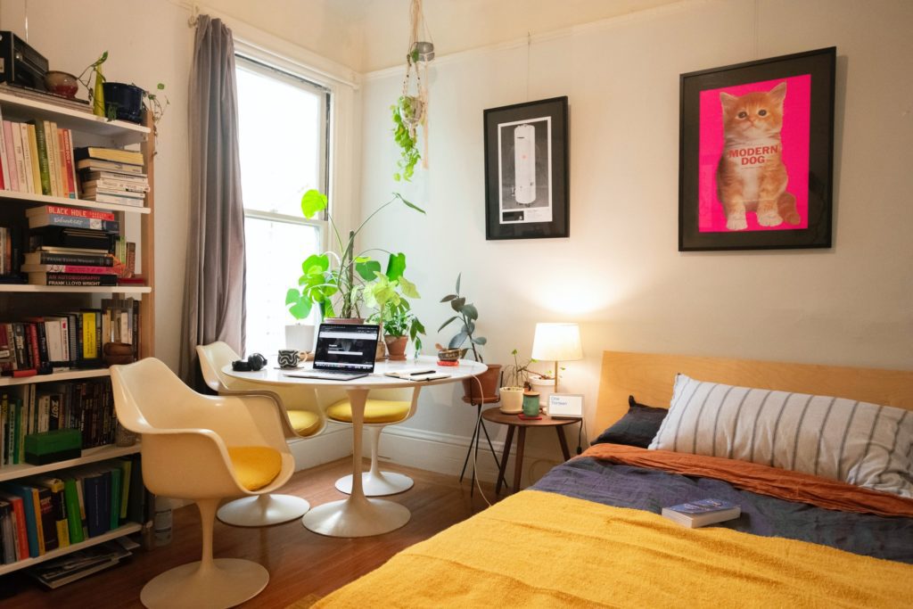 Bedroom with a bed with a yellow blanket, bookcase, round white desk and yellow chair. 