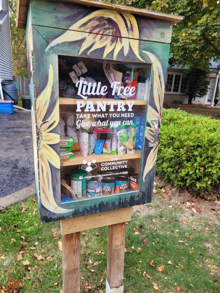 A wooden cupboard with four shelves filled with food having a glass door mounted on a wooden stand